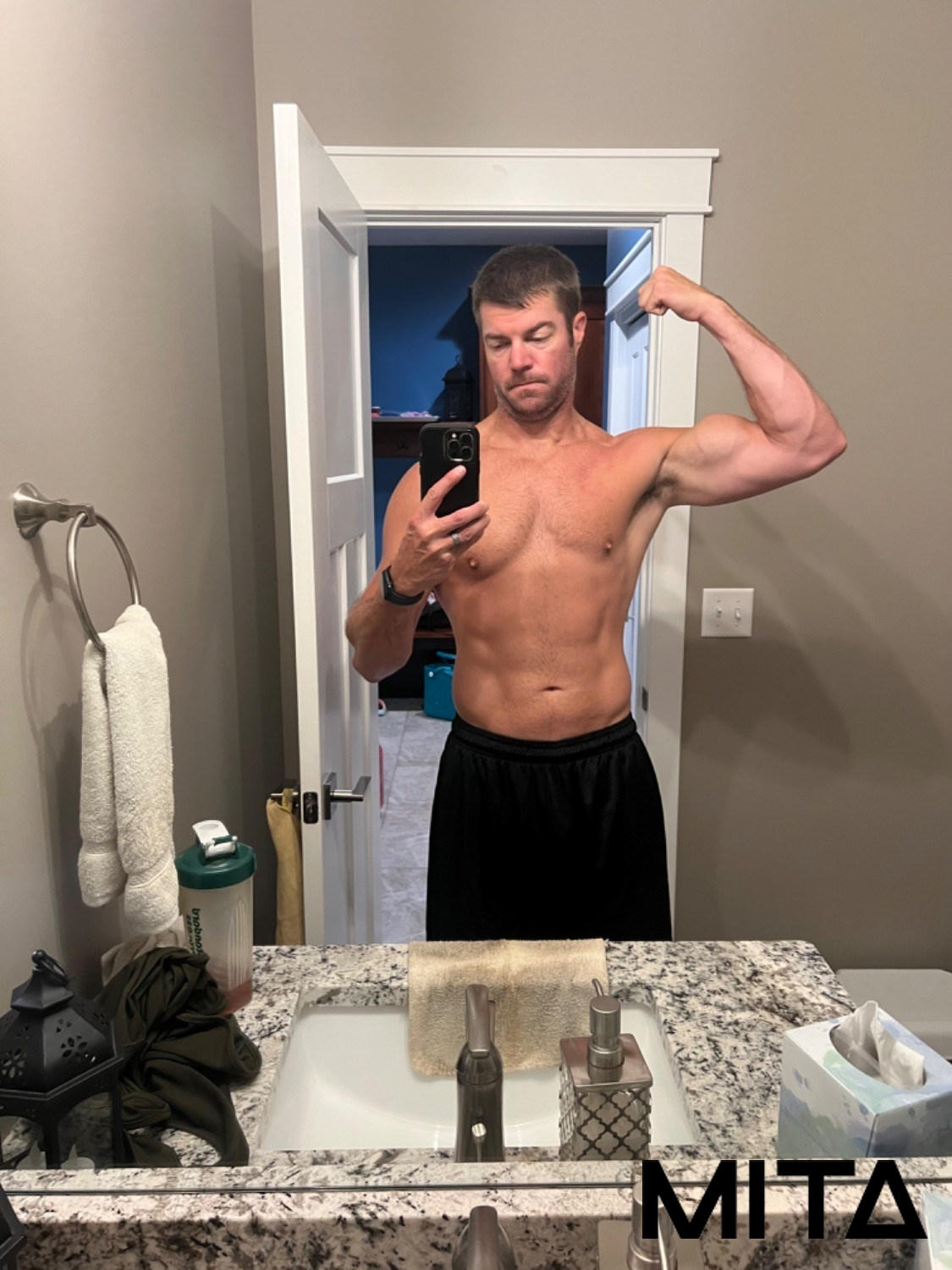 Man taking a shirtless mirror selfie, flexing his bicep in a bathroom.