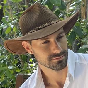 Man wearing a brown hat and white shirt, surrounded by greenery.